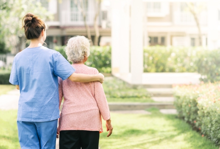 A caregiver in blue scrubs places her arm over the shoulder of an elderly woman as they walk together outside.