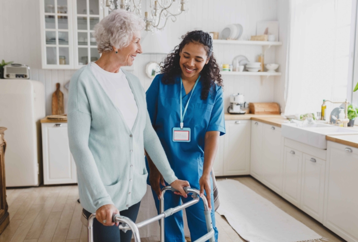 Medical nurse in home care helping senior female at home, teaching her to walk with walker, smiling and giving instructions how to use device, standing together in cozy kitchen.