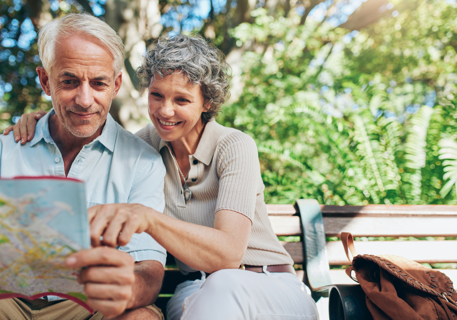Portrait of happy man and woman reading map while sitting on a park bench. Senior couple on vacation using city map for travel tips for seniors.
