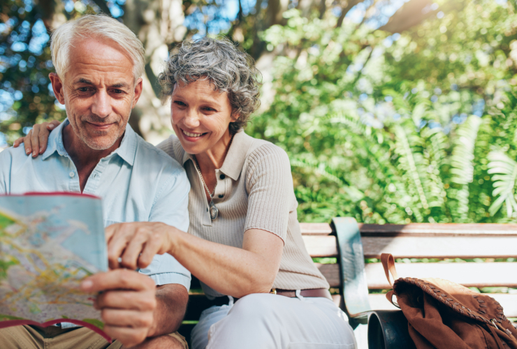 Portrait of happy man and woman reading map while sitting on a park bench. Senior couple on vacation using city map for travel tips for seniors.