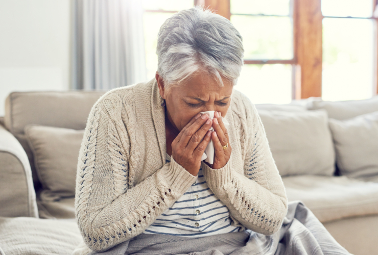 Senior woman, sick during flu season and blowing nose at home from virus, allergy and illness on couch. Tired, tissue and elderly female person in a house lounge with sneeze from covid and flu on a sofa with allergies.